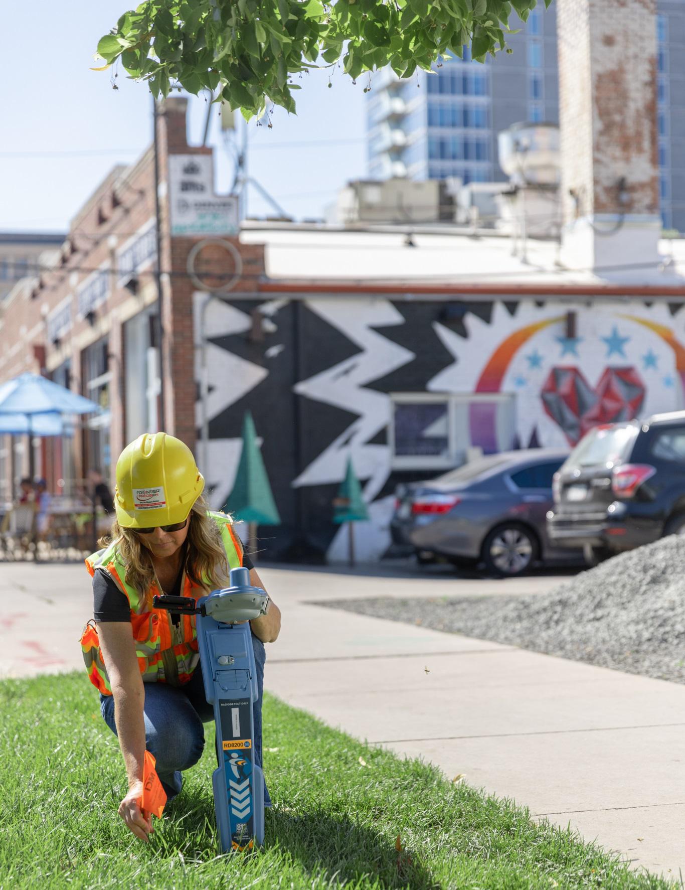 A woman surveying, in a high-visibility jacket and hard hat, kneeling on a sunny street